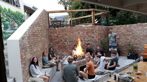 A group of people sitting around a fire pit in an outdoor patio area with brick walls and a wooden deck. - Island Vibe Knysna Hostel (Knysna)