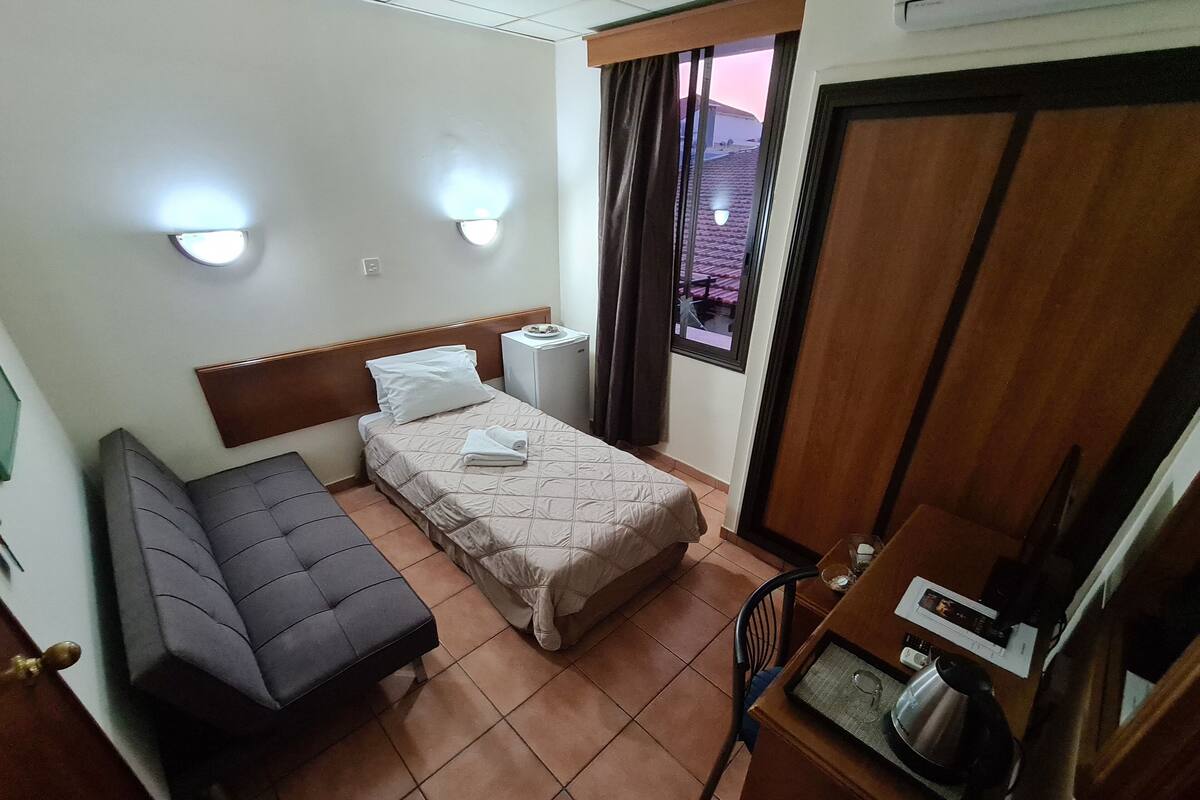 A hotel room with a bed, a sofa, a small table with a kettle, and a window with curtains.