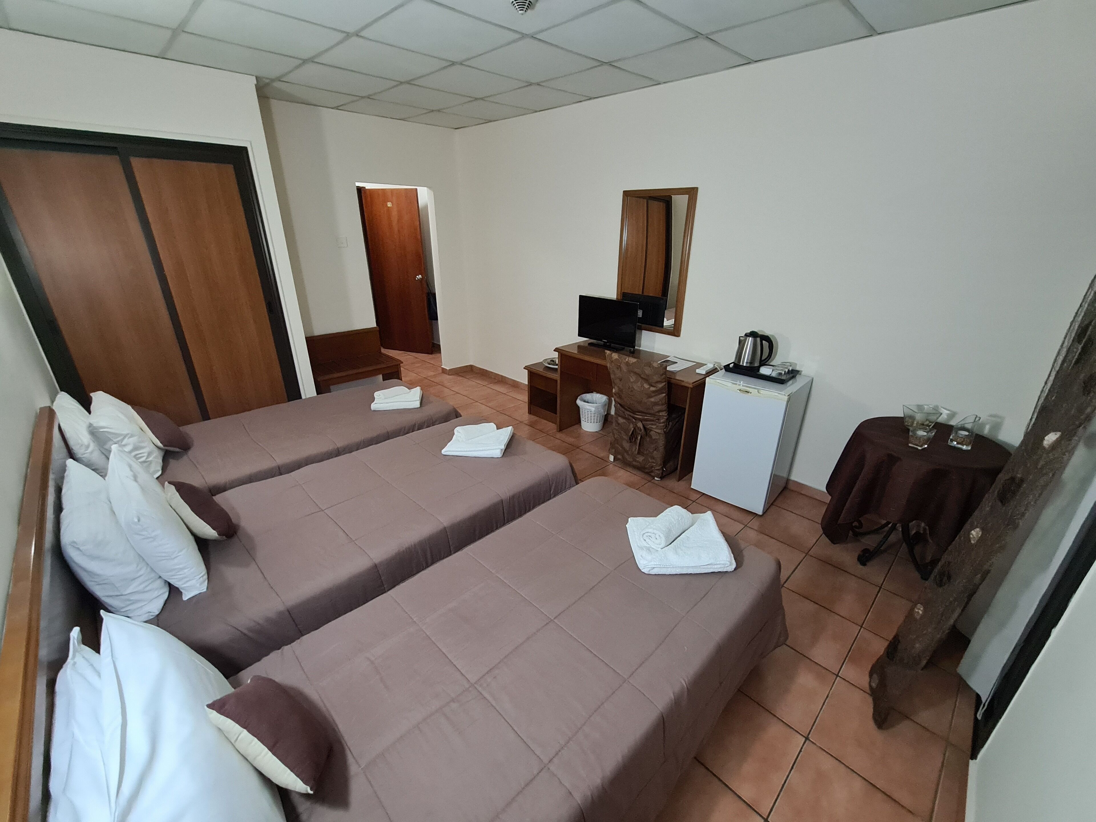 A hotel room with two beds, a desk with a computer, a small refrigerator, and a table with glasses.