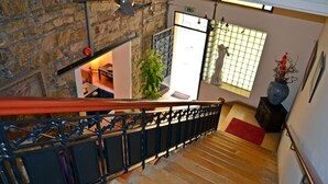 A staircase with a wooden handrail, a stone wall, and a view of a room with a plant and a red rug. - Metropole Hotel (Limassol)