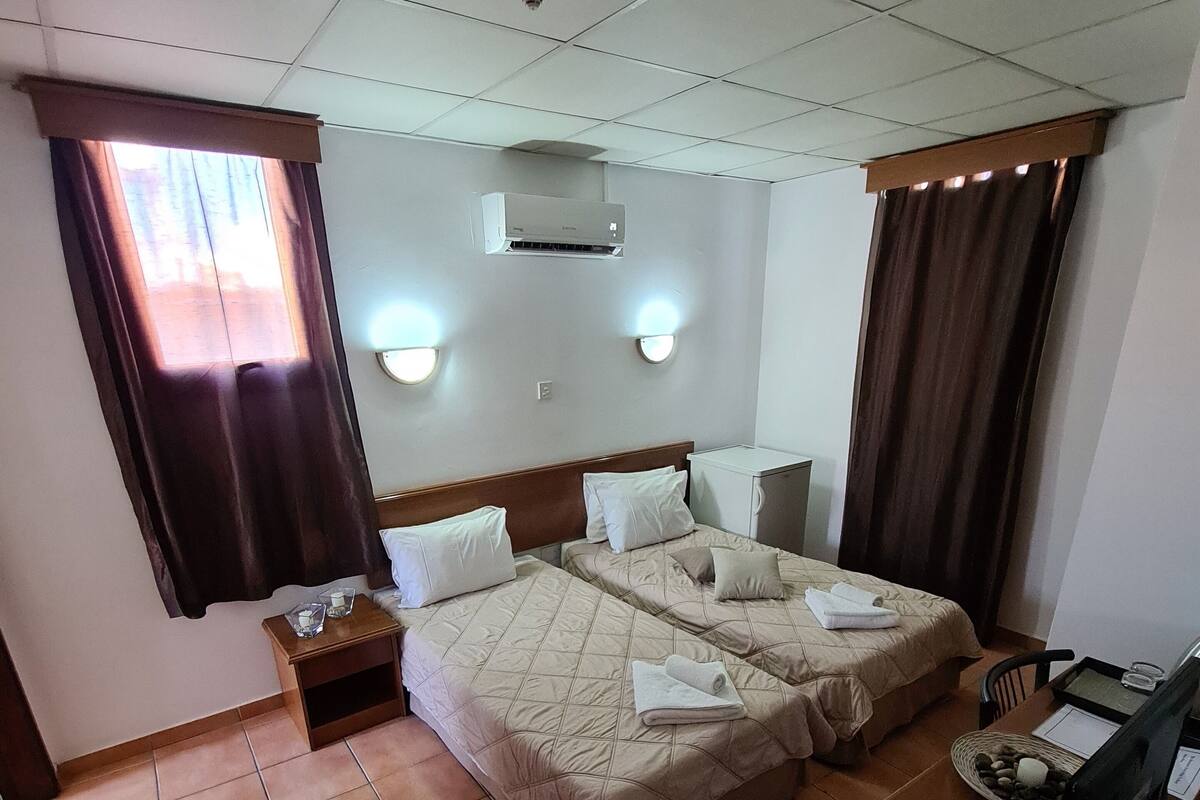 A hotel room with two beds, a desk, a chair, a small table, a nightstand, a window with curtains, wall-mounted lights, and an air conditioning unit.