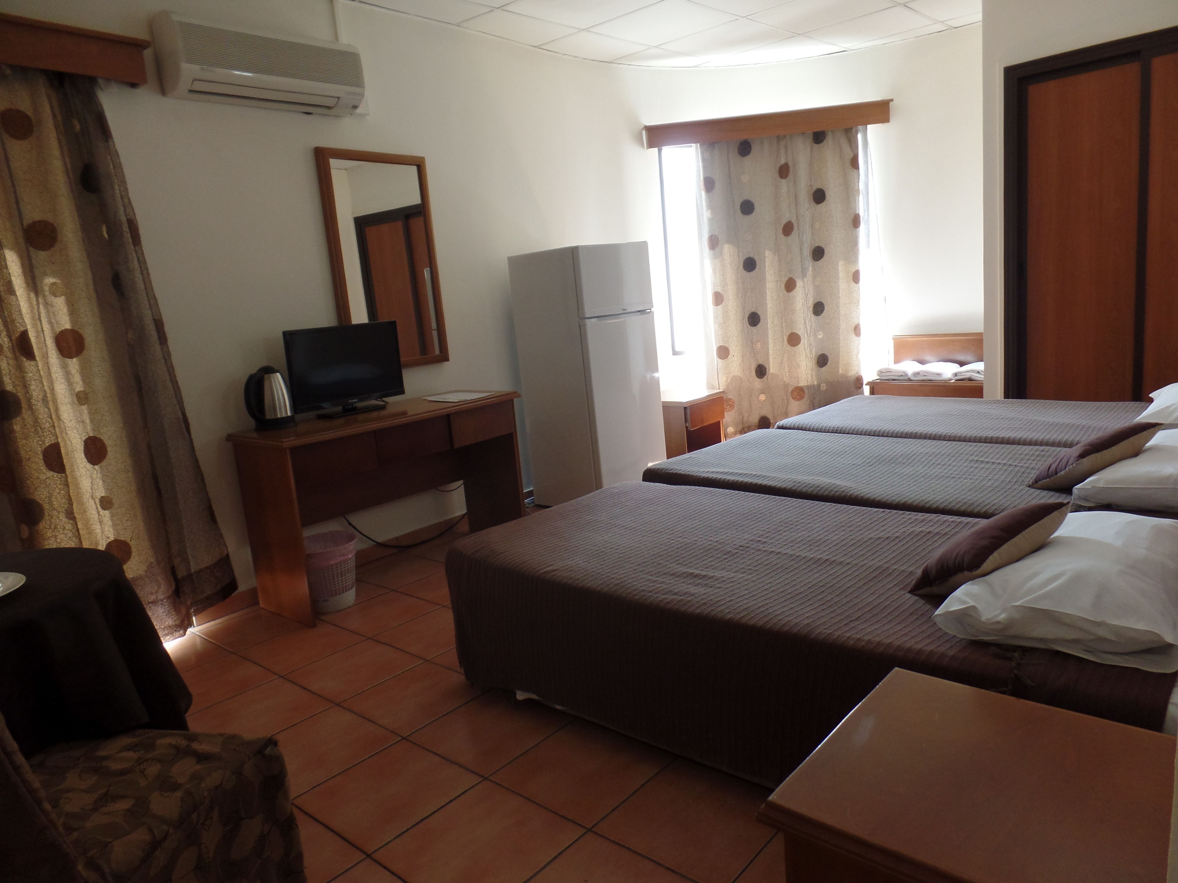 A hotel room with two beds, a desk with a television, a refrigerator, and a window with curtains.