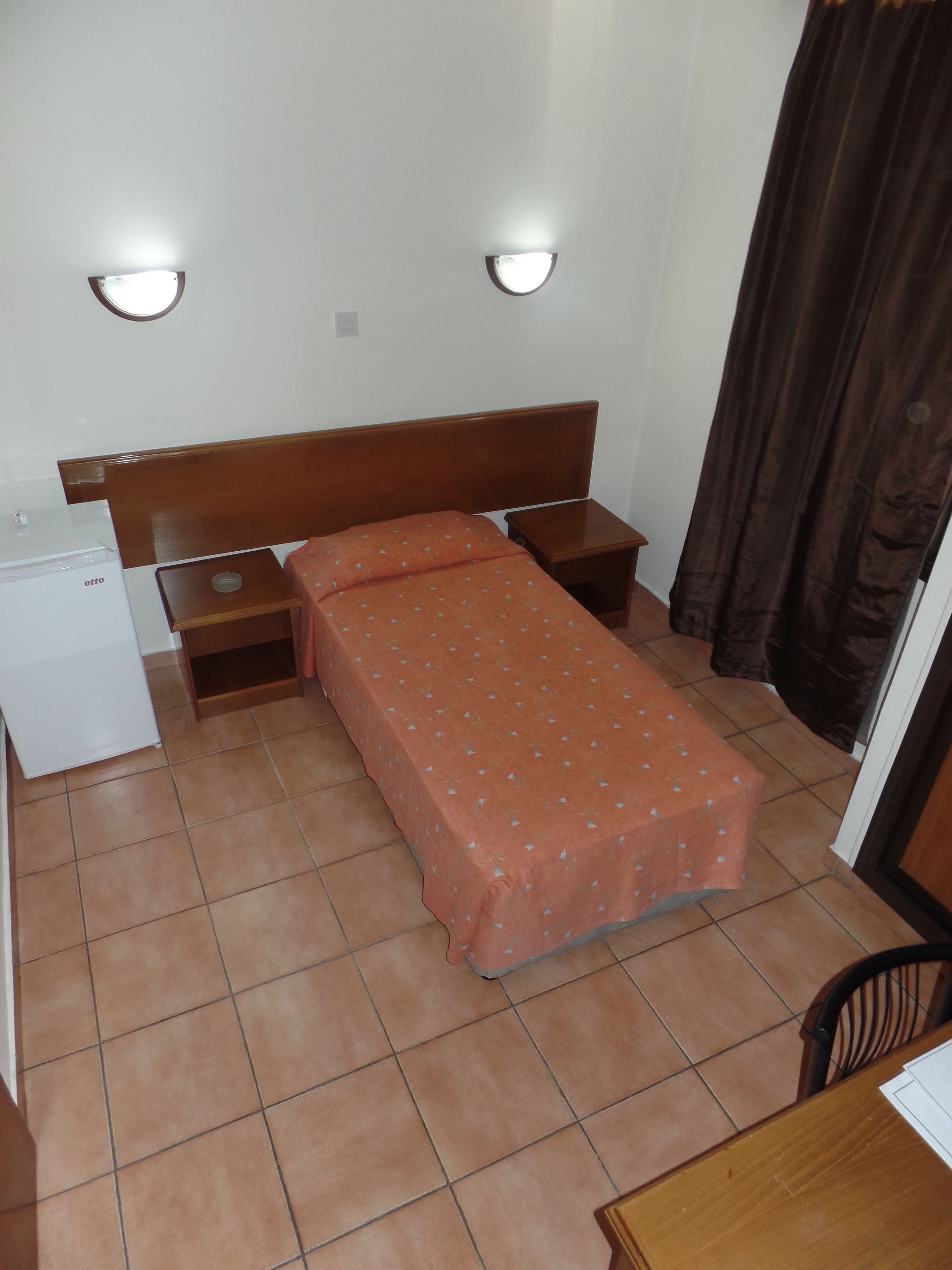 Een kleine, eenvoudige kamer met een bed, een nachtkastje, een bureau en een koelkast.