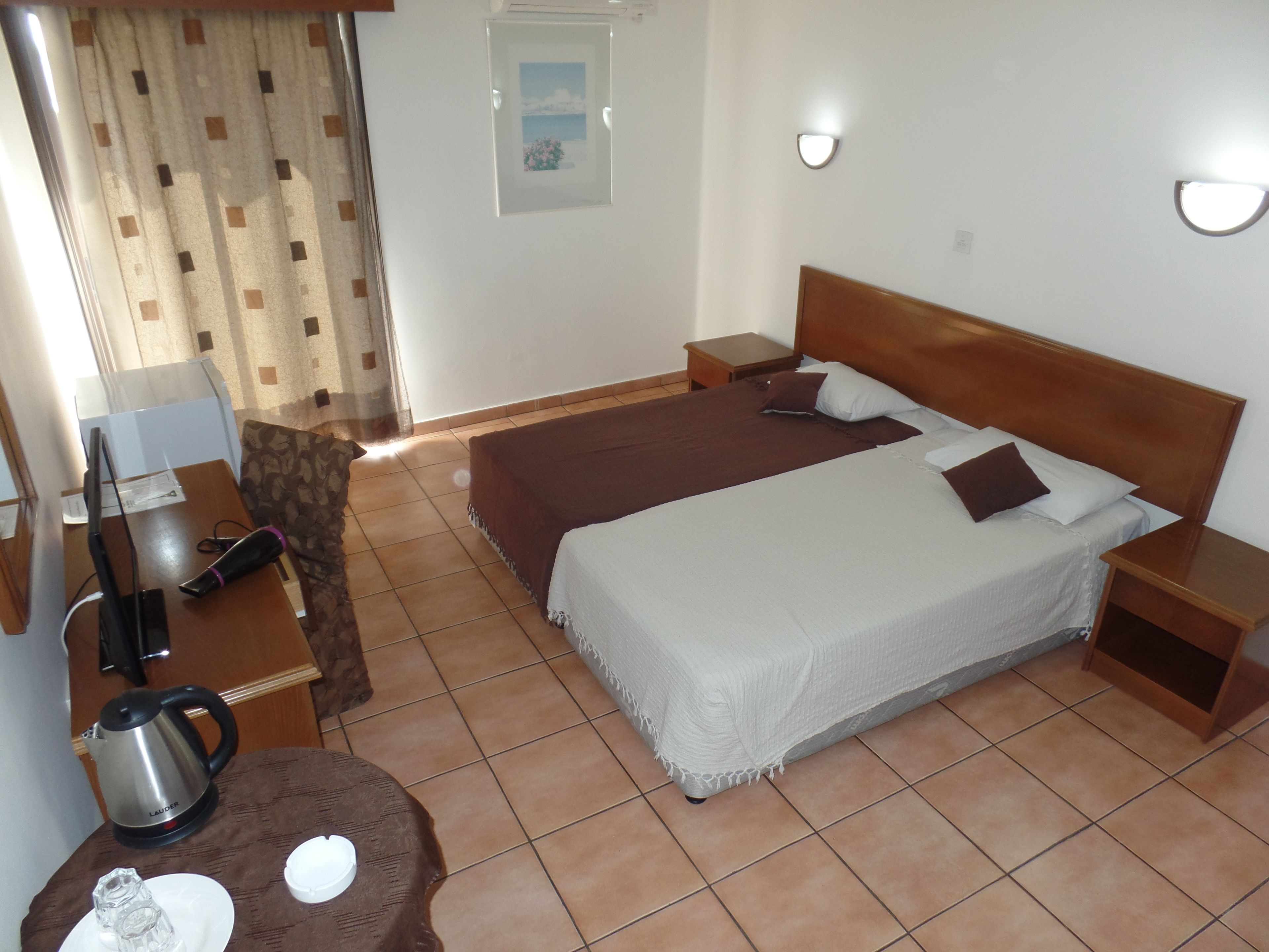 Une chambre d’hôtel comprenant un lit, des tables de chevet, un bureau avec une bouilloire, une chaise et une fenêtre avec des rideaux.