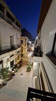 A narrow street with buildings on either side, outdoor seating areas, and a few parked cars.