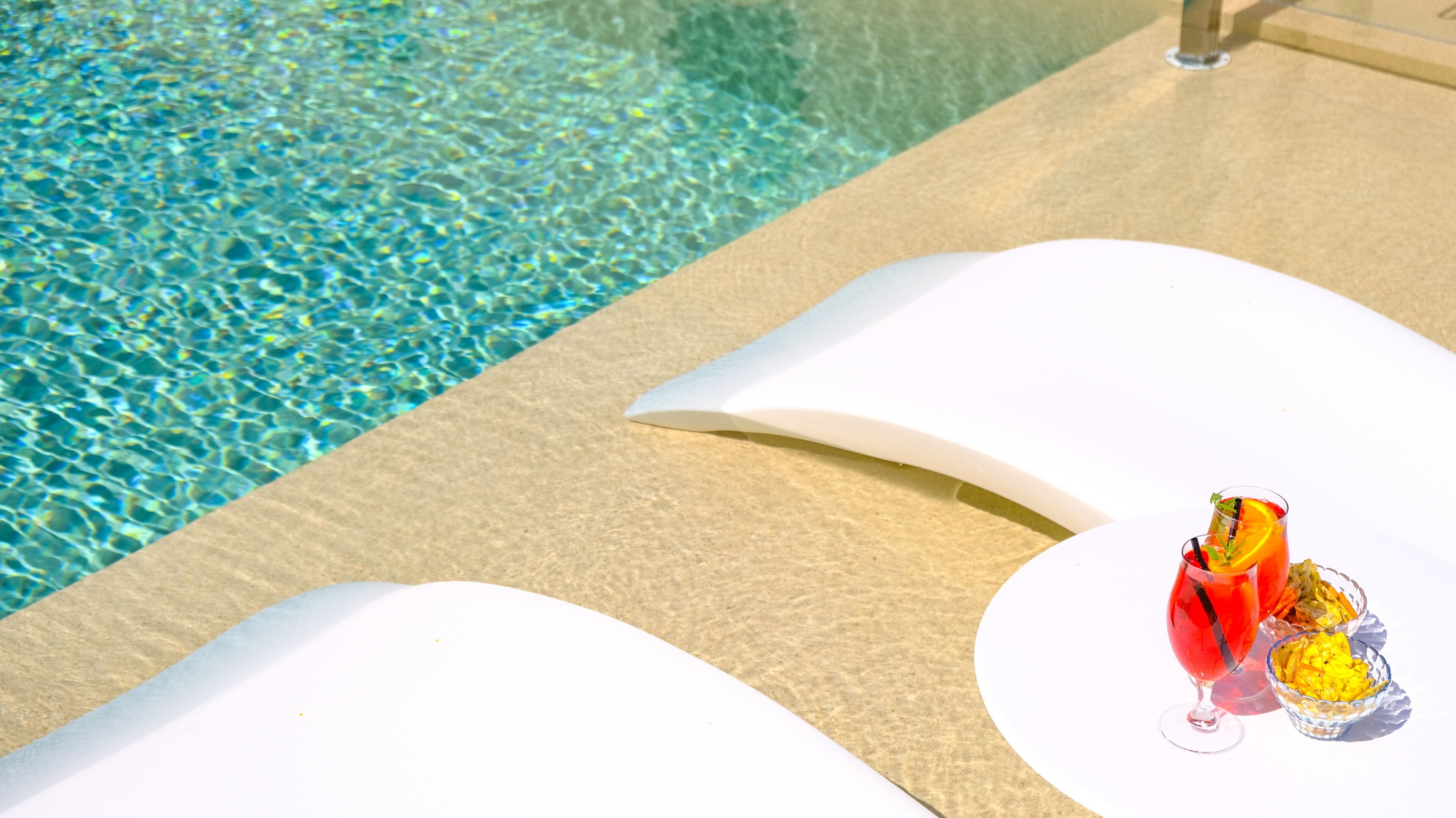 A poolside area with clear water and a lounge chair. There are two glasses of red drink with garnishes and a bowl of yellow snacks on the chair.