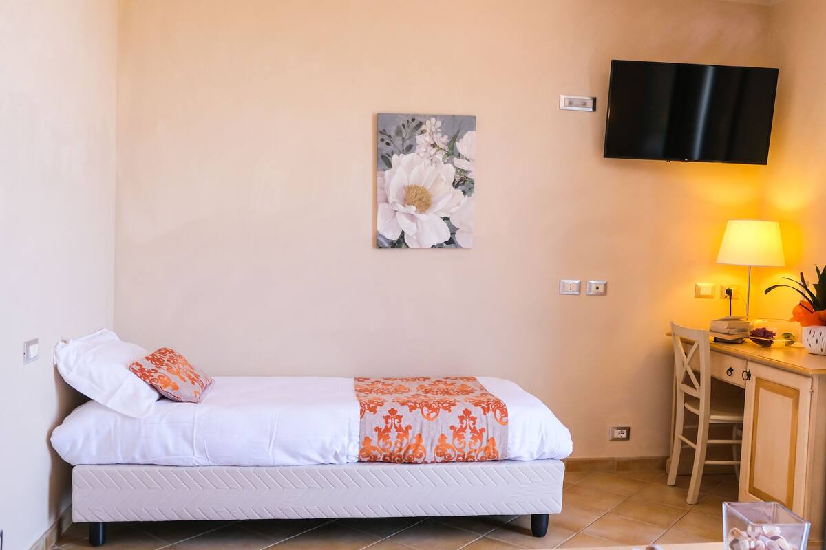 A single bed with a white and orange bedspread, a mounted TV, a bedside lamp, and a small table with a lamp and decorative items.