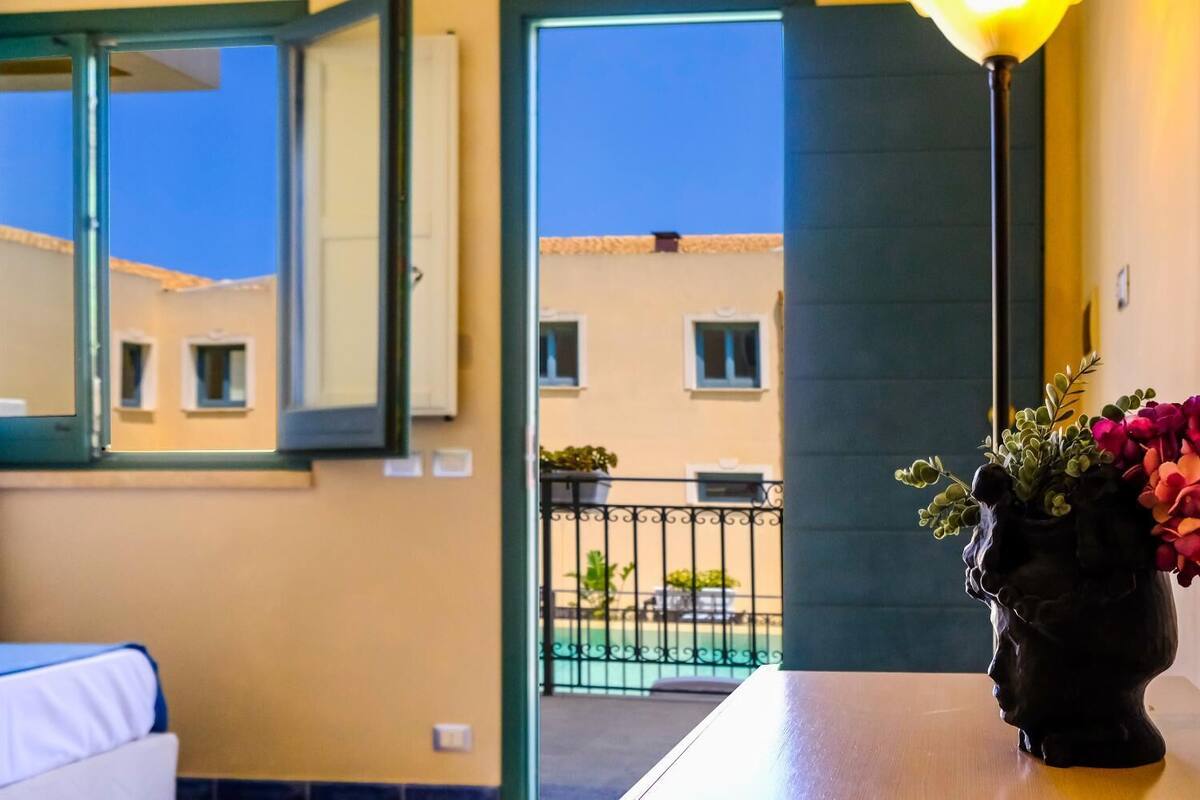 A room with a view of a building through an open window, a table with a vase of flowers, and a lamp.