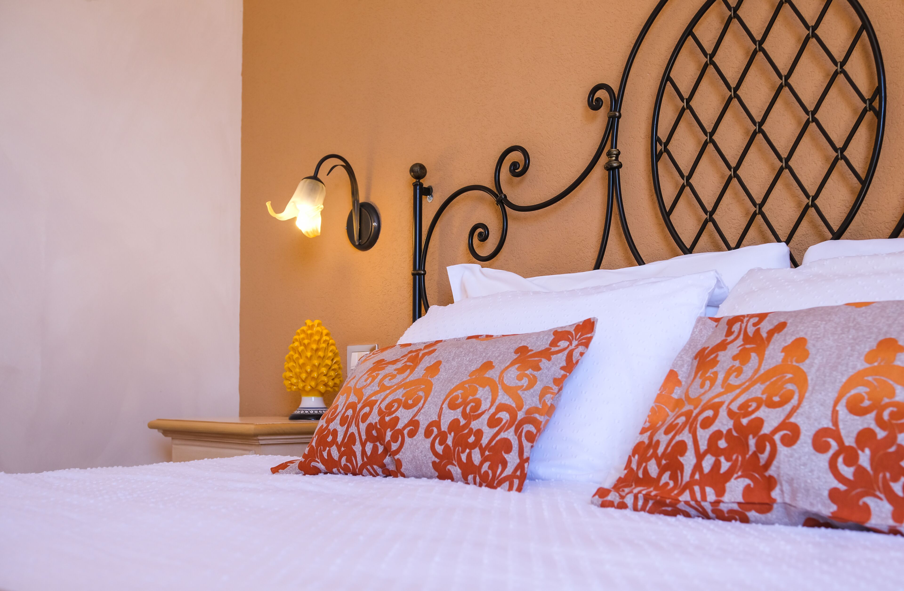Uma cama bem arrumada com almofadas brancas e laranja estampadas, um painel de parede decorativo e uma luminária fixada na parede.