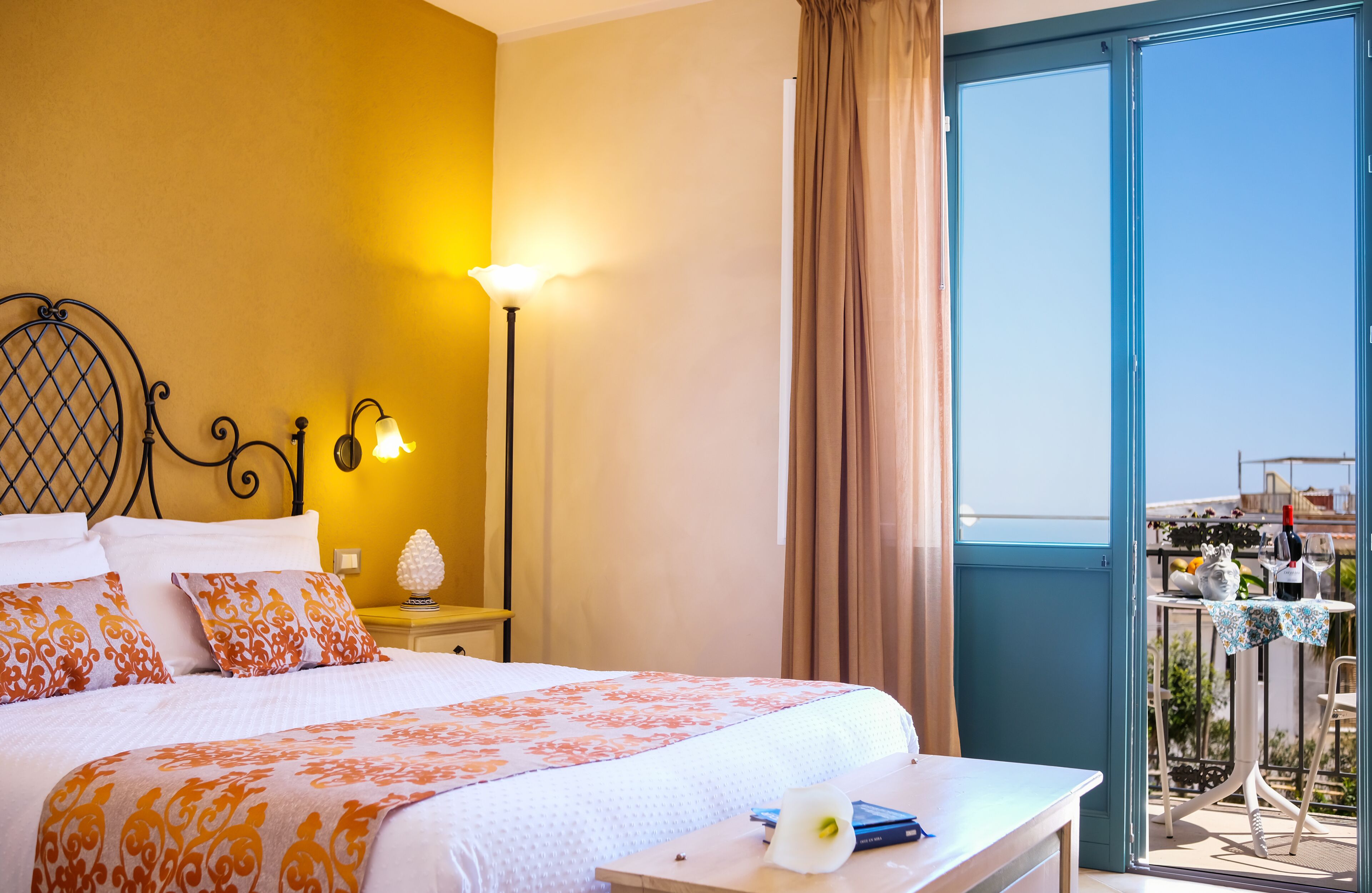 Habitación de hotel con cama, lámpara de noche y vistas al balcón con mesa y sillas.