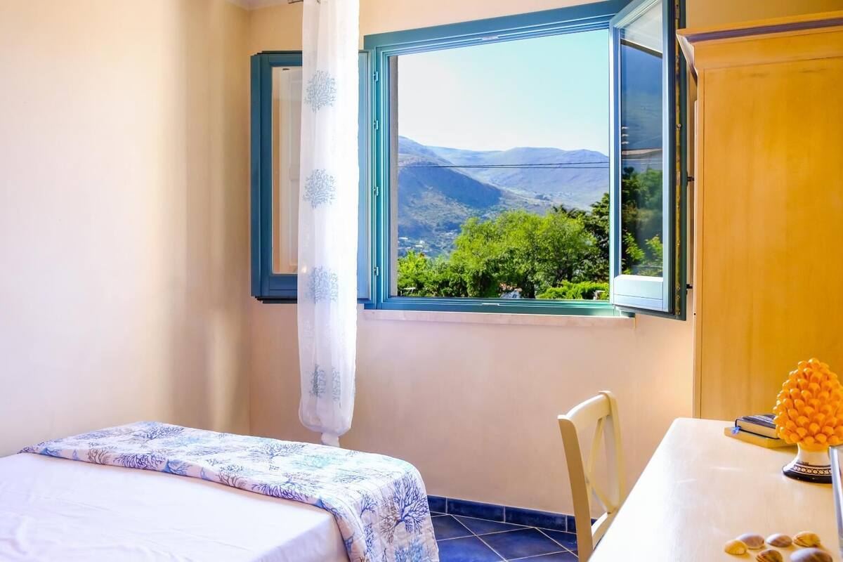 A bedroom with a bed, a chair, a table, and a window with a view of mountains and greenery.