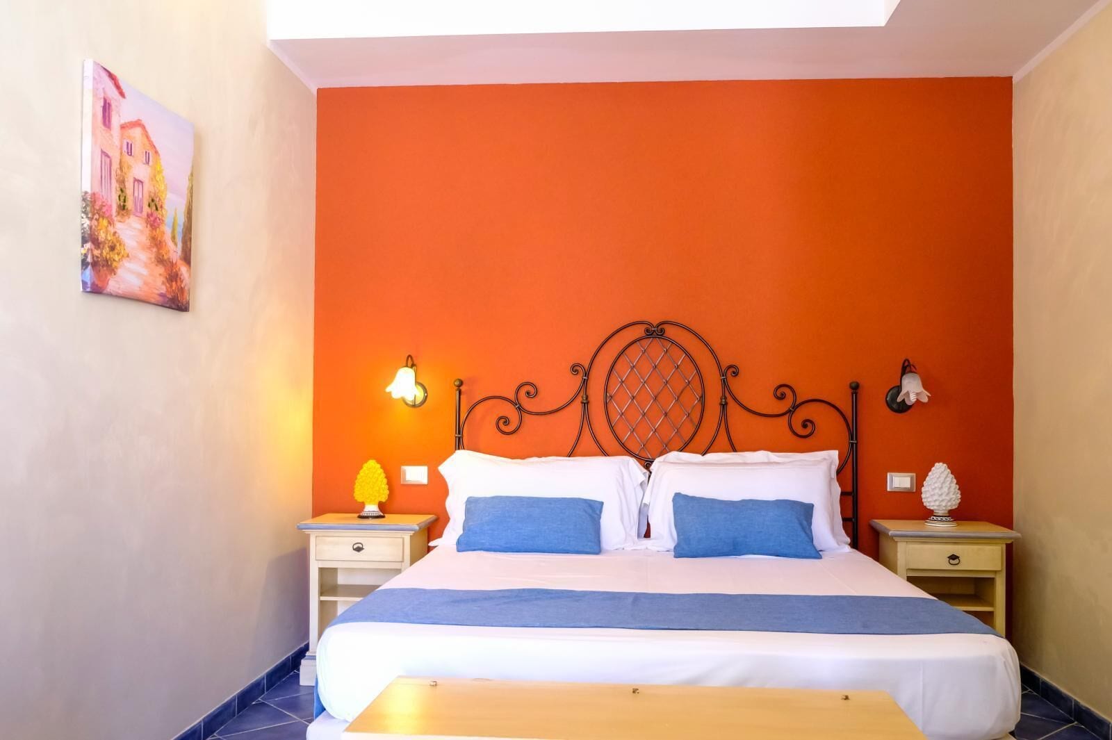 A bedroom with an orange wall, a bed with white and blue bedding, two bedside tables with lamps, and a painting on the wall.