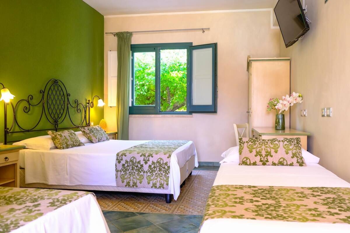 A hotel room with two beds, a large window with a view of greenery, a bedside table with a lamp, and a TV mounted on the wall.