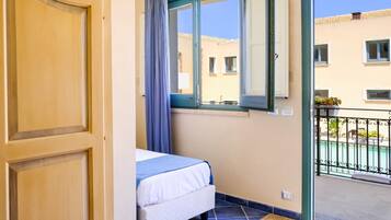 A room with a bed, a window with a view of buildings, and a balcony with a railing.