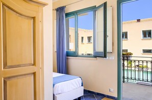 A room with a bed, a window with a view of buildings, and a balcony with a railing. - Hotel Baglio di Scopello (Castellammare del Golfo)