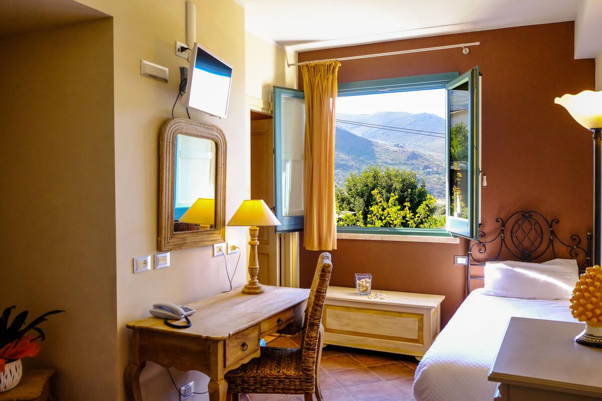 A hotel room with a desk, chair, bed, and a window with a view of greenery and mountains.