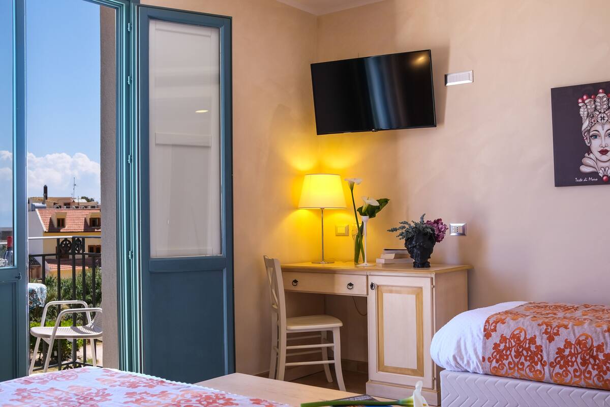 A hotel room with a bed, a desk with a chair, a TV mounted on the wall, and a window with a view of buildings.
