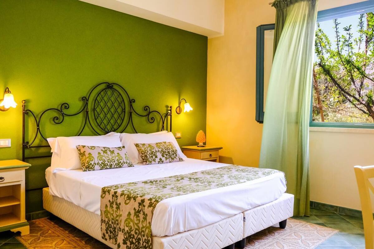 A neatly made bed with white linens and green patterned pillows, a green wall, and a window with a view of trees.