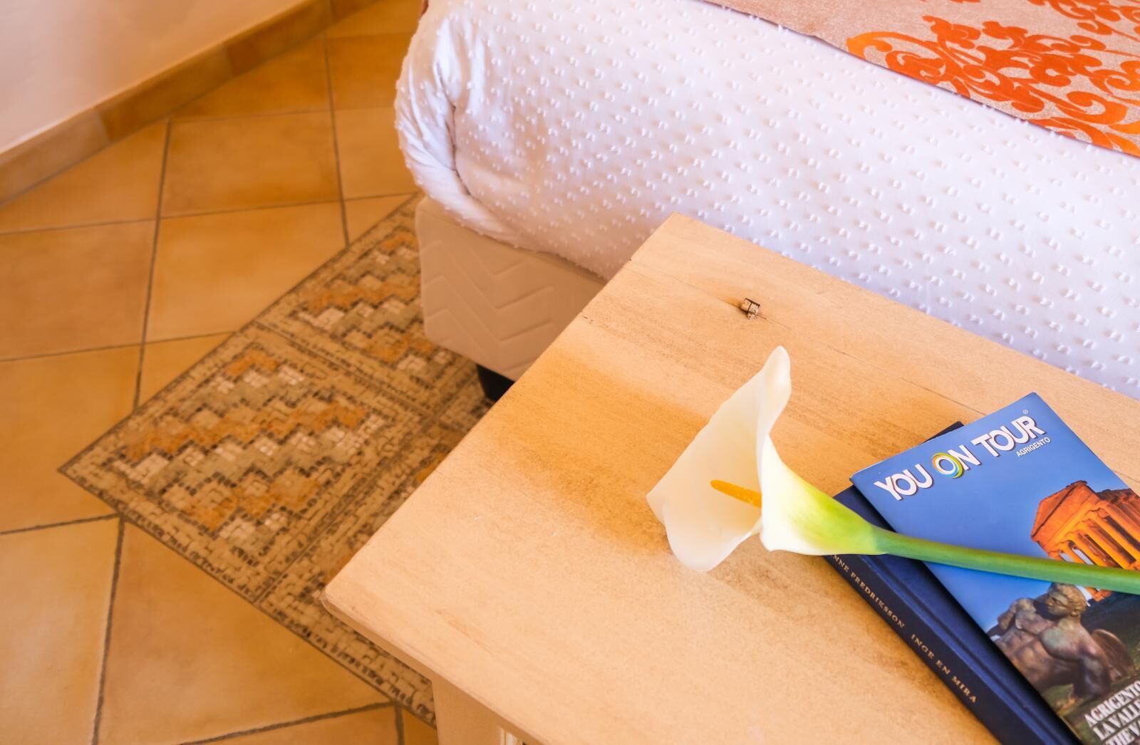 Un lit recouvert d’une couverture blanche à texture, une table de chevet en bois sur laquelle repose un livre intitulé « Vous en tournée » et une fleur blanche.