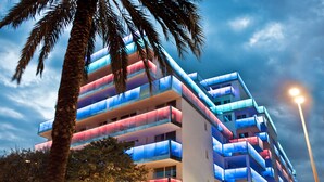 Een meerlaags hotel met balkons die verlicht zijn in blauw en rood, omringd door palmbomen.
