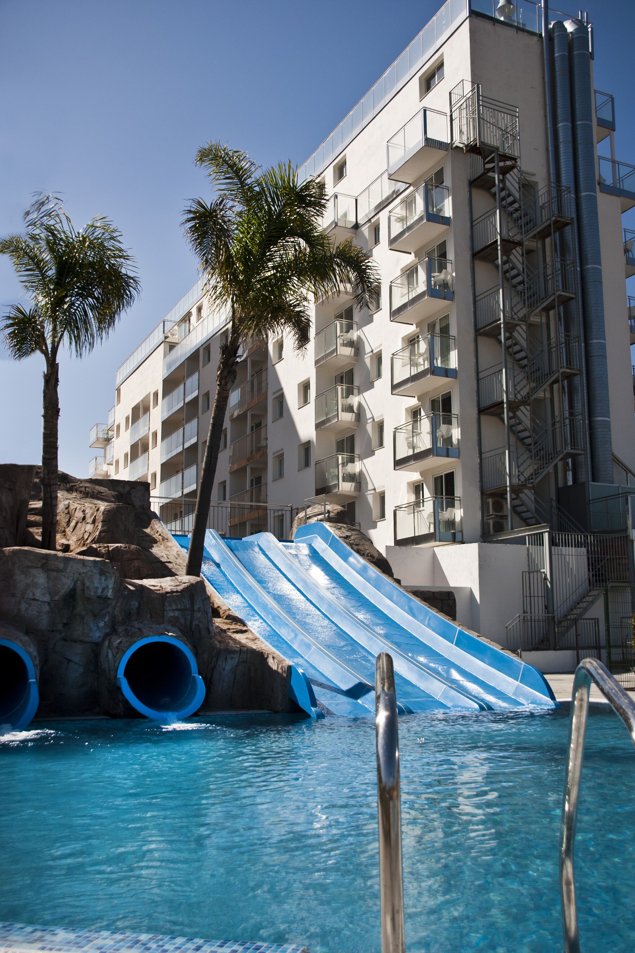Une piscine avec un toboggan bleu, des palmiers et un immeuble à plusieurs étages doté de balcons et d’un escalier.