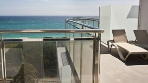 Een balkon met glazen hekwerk met uitzicht op de oceaan, twee ligbedden en een witte constructie.