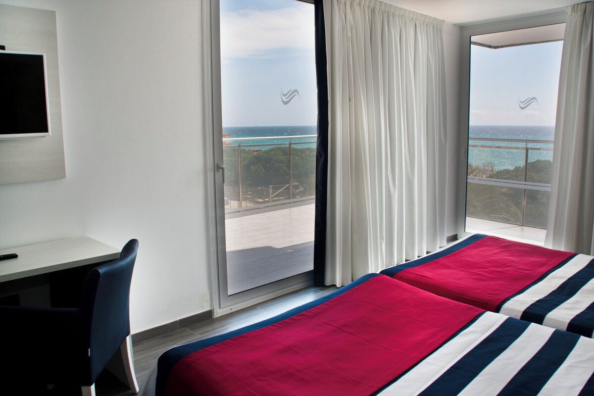Habitación de hotel con cama, escritorio y vista al océano a través de un ventanal grande.