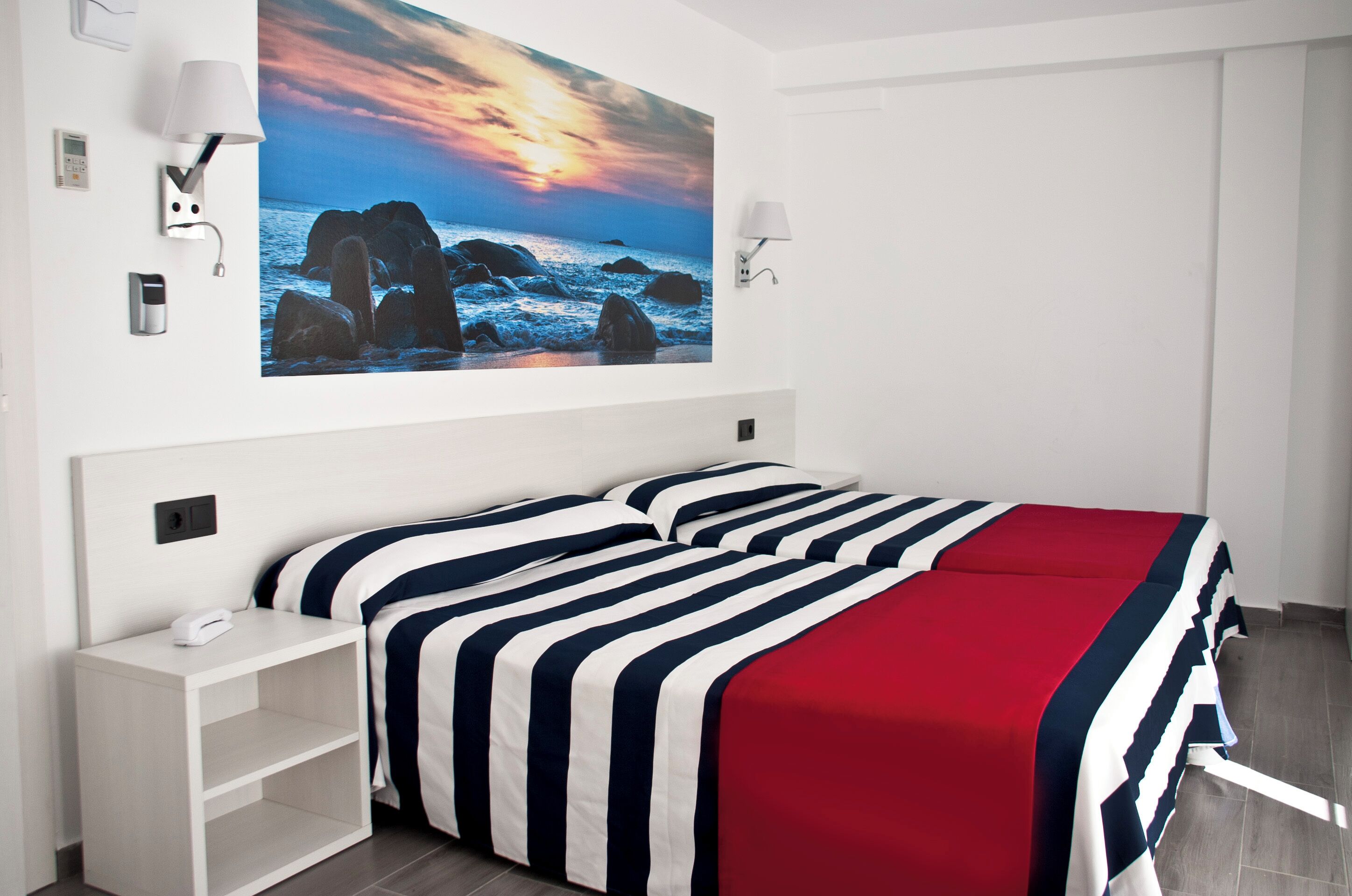 Une chambre d’hôtel avec un lit, une table de chevet, une lampe fixée au mur et un grand tableau représentant un coucher de soleil sur la mer.