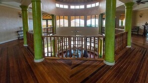 A circular room with a wooden floor and green pillars, featuring a central wooden railing and a view of the outdoors through windows. - Milbrooks Villa and Retreat (Montego Bay)