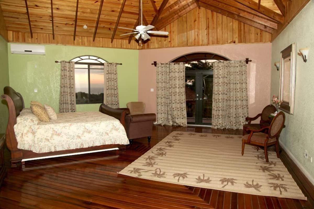 A bedroom with a wooden ceiling, a bed, two chairs, a rug, and a window with curtains.