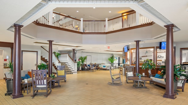 A spacious lobby with a wooden herringbone floor, a central seating area with sofas and chairs, a staircase connecting two levels, and a large plant in the center. - Oakwood Resort (Syracuse)