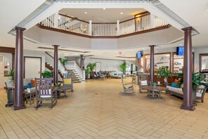 A spacious lobby with a wooden herringbone floor, a central seating area with sofas and chairs, a staircase connecting two levels, and a large plant in the center. - Oakwood Resort (Syracuse)