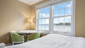 A hotel room with a bed, a small round table, two chairs, a lamp, and a large window with a view of a body of water. - Oakwood Resort (Syracuse)