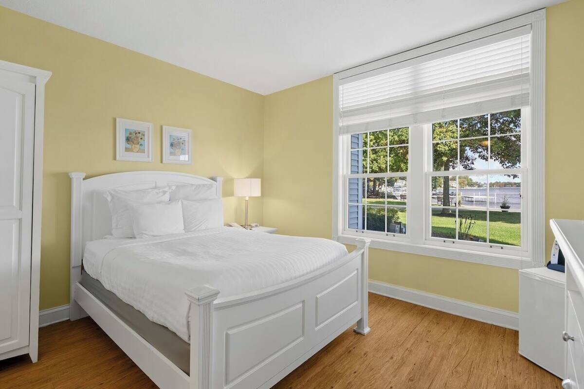 A bedroom with a bed, a nightstand, a lamp, a window with a view of the outdoors, and a door.