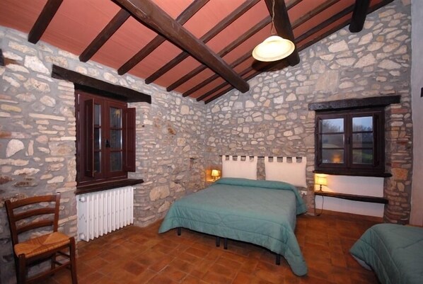 A bedroom with stone walls, a wooden ceiling, a bed with a teal bedspread, two bedside lamps, and a chair. - Agriturismo Le Casette (Montecchio)