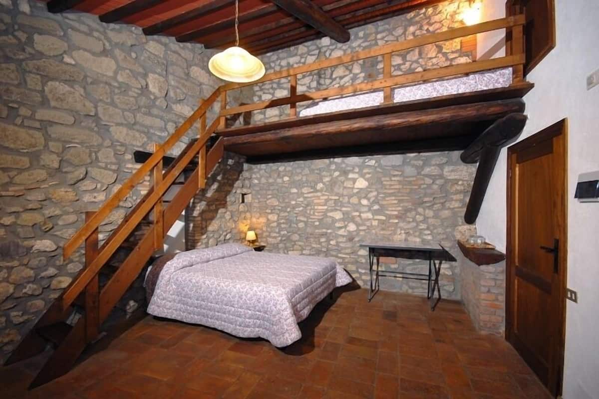 Un dormitorio con pared de piedra, escalera de madera, cama con manta estampada, mesa con lámpara y una pequeña estantería.