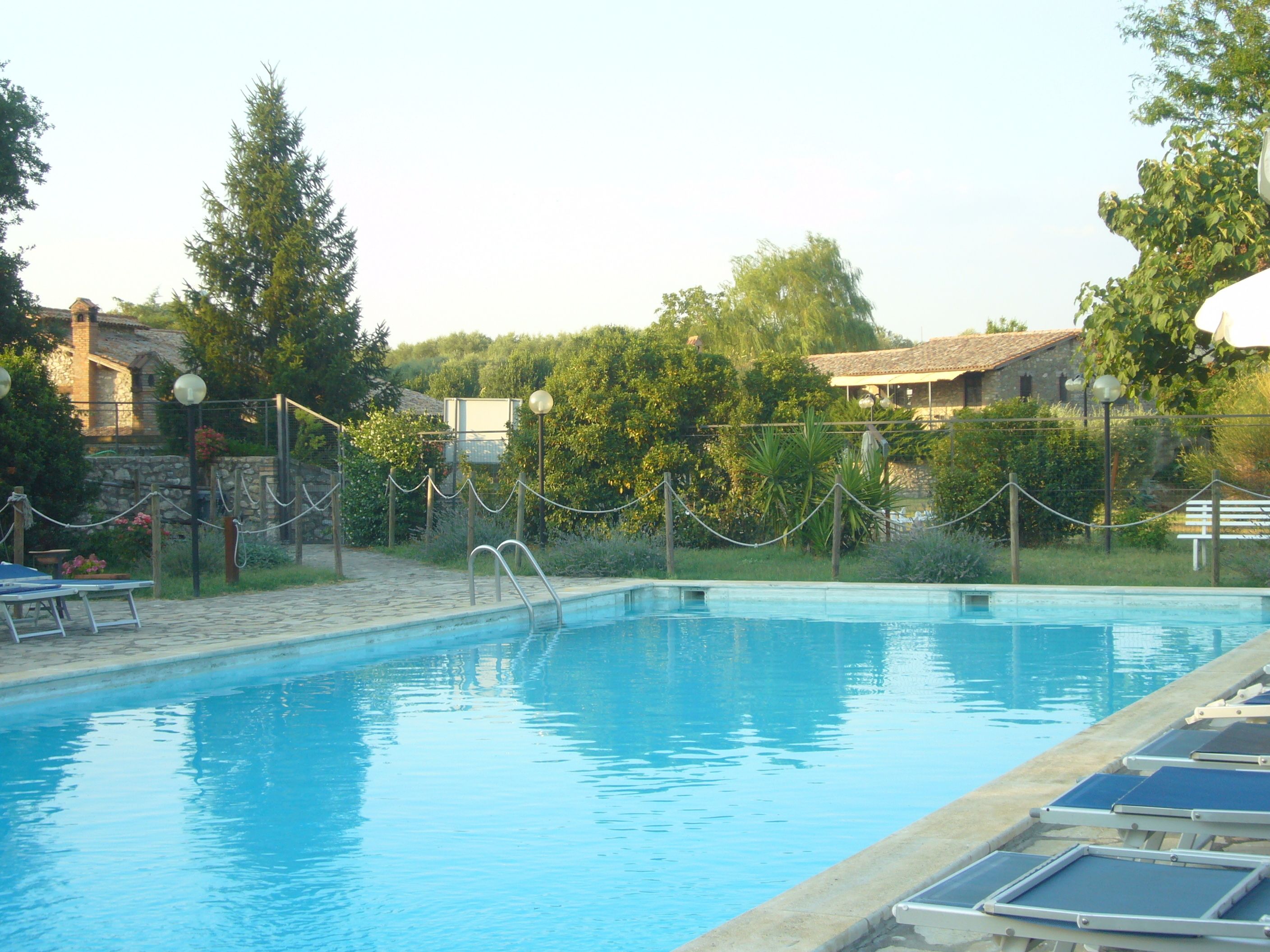 A swimming pool with a clear blue water, surrounded by a stone deck and a wooden fence. There are lounge chairs along the poolside.