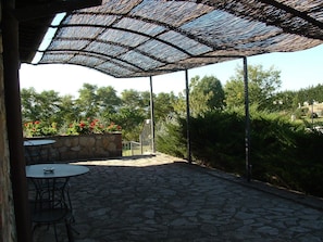 Un patio al aire libre con suelo de piedra, sillas de metal y una estructura de sombra tejida.