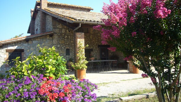 Un edificio de piedra con techo de tejas, un patio con mobiliario exterior y plantas en flor.