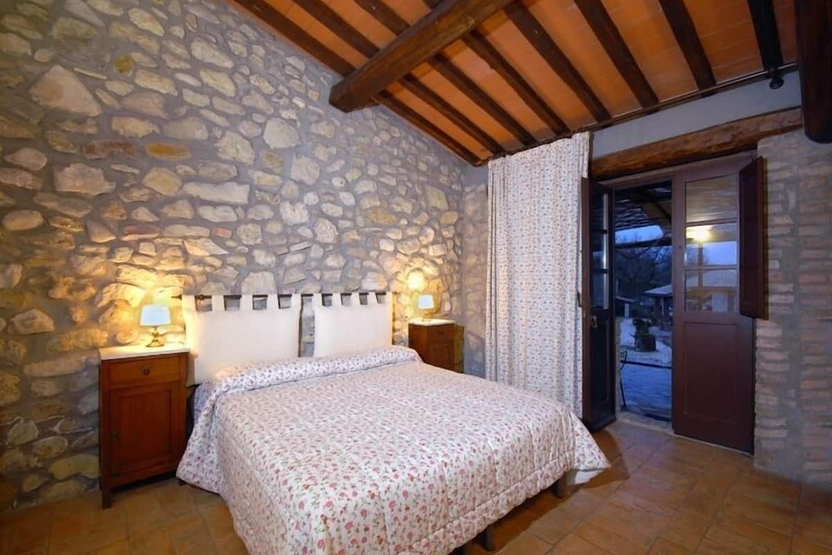 Un dormitorio con pared de piedra, vigas de madera en el techo, una cama con colcha estampada, dos lámparas de noche y una puerta que da al exterior.