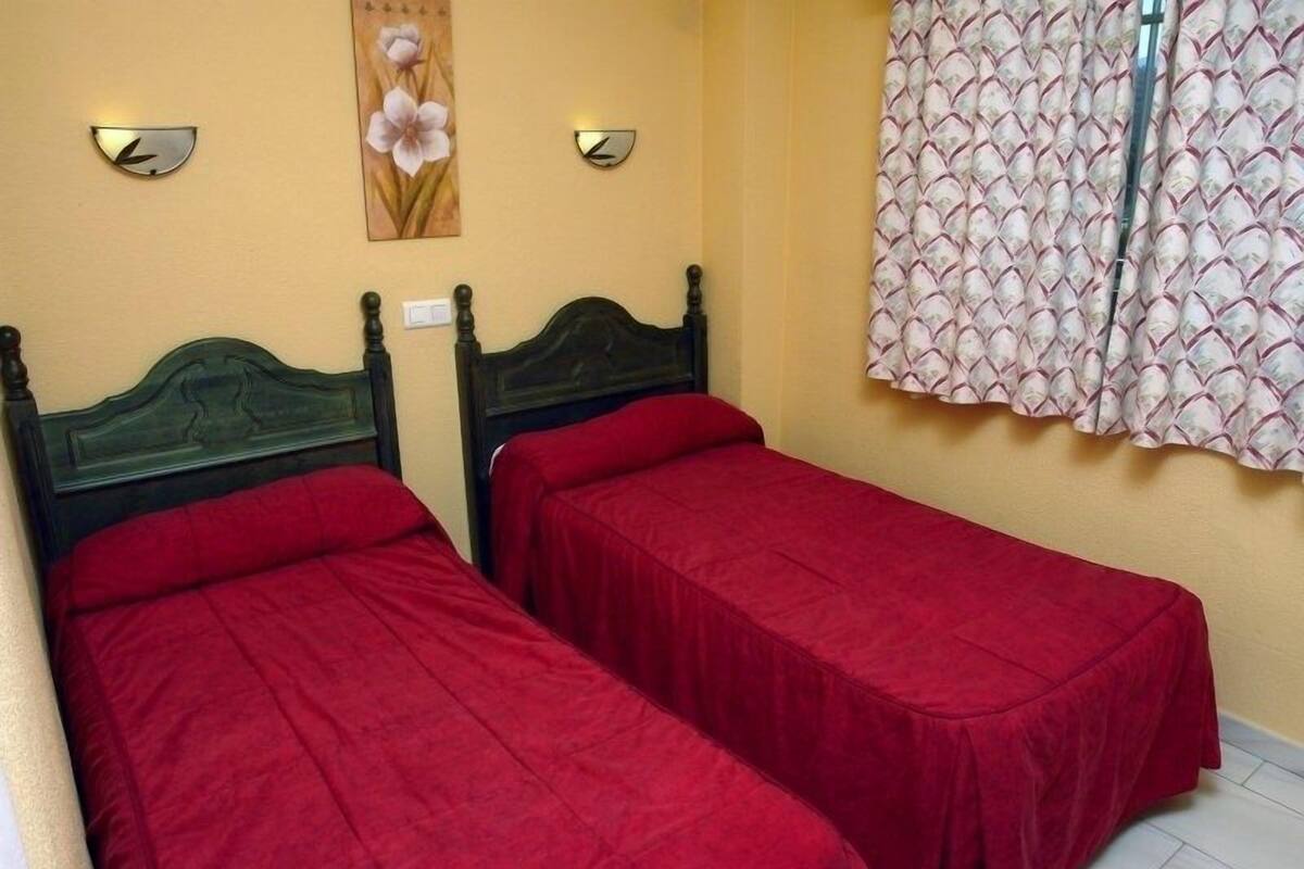 A room with two single beds, each with a red bedspread, a wooden headboard, and a wall-mounted light fixture.
