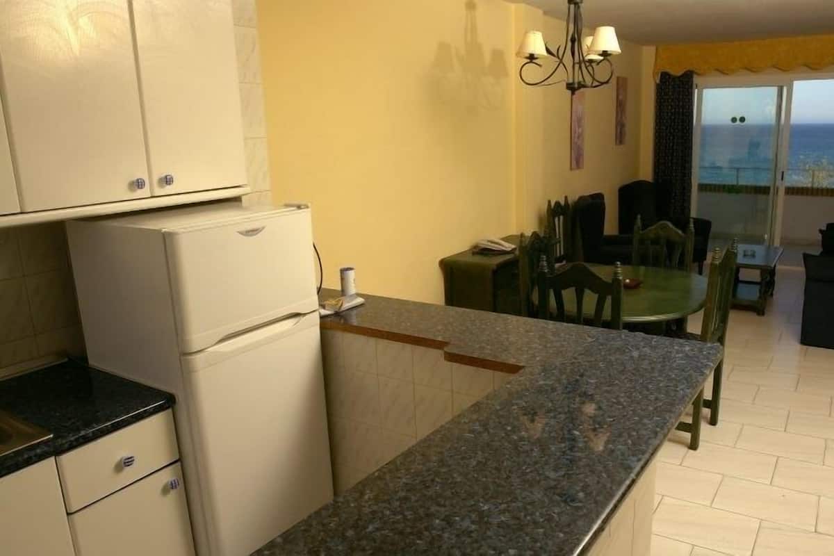 A compact kitchen with a refrigerator, a dining table with chairs, and a view of the sea through a window.