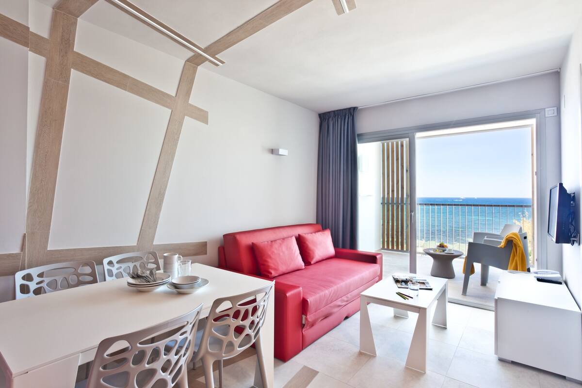 Habitación de hotel moderna con sofá rojo, mesa de comedor blanca y balcón con vistas al mar.