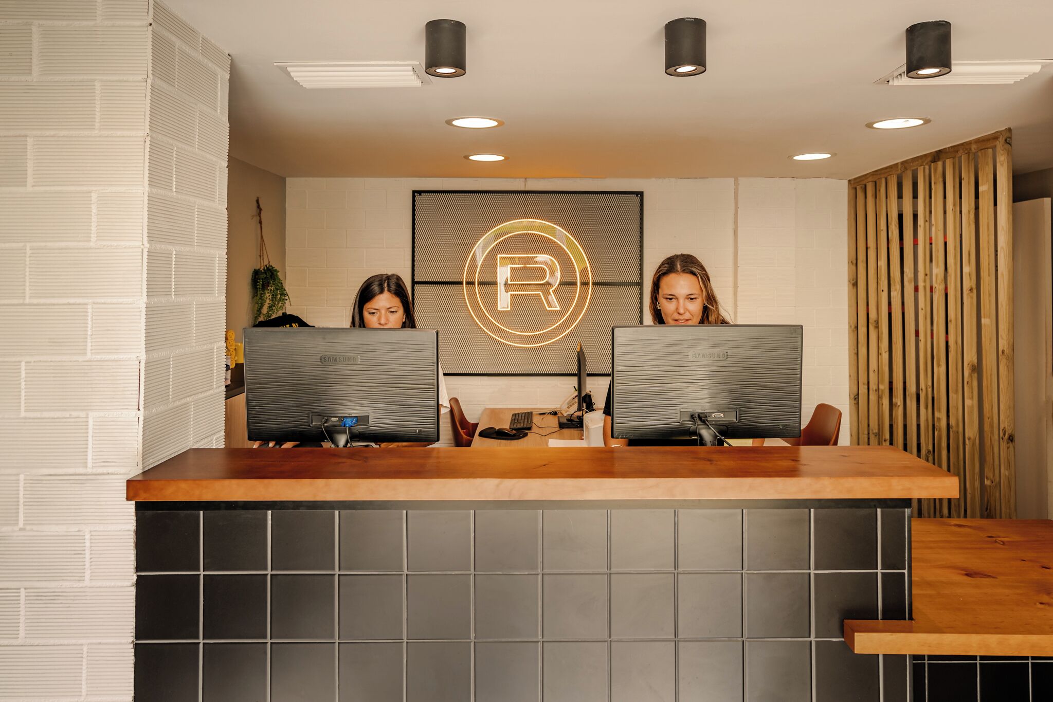 A reception desk with two computer monitors, a logo on the wall, and a wooden panel on the right.