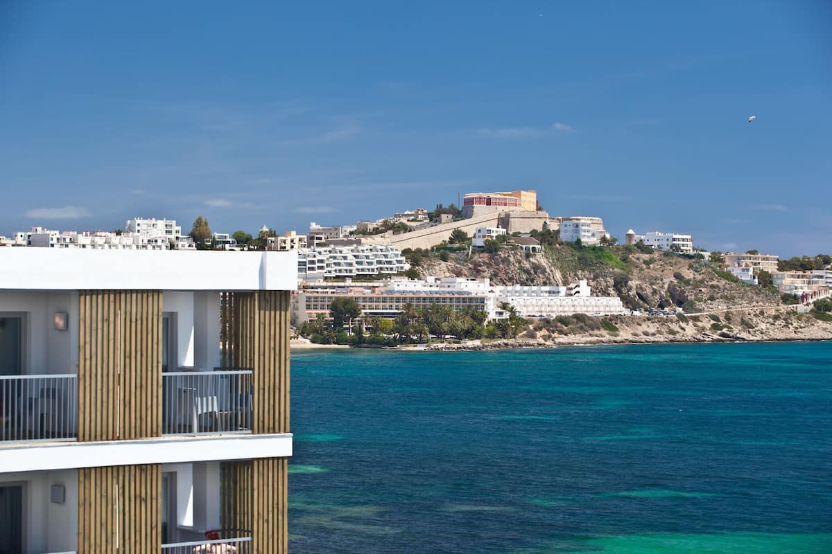 Un hotel frente al mar con varios pisos, balcones y vistas al océano y a una colina con edificios.