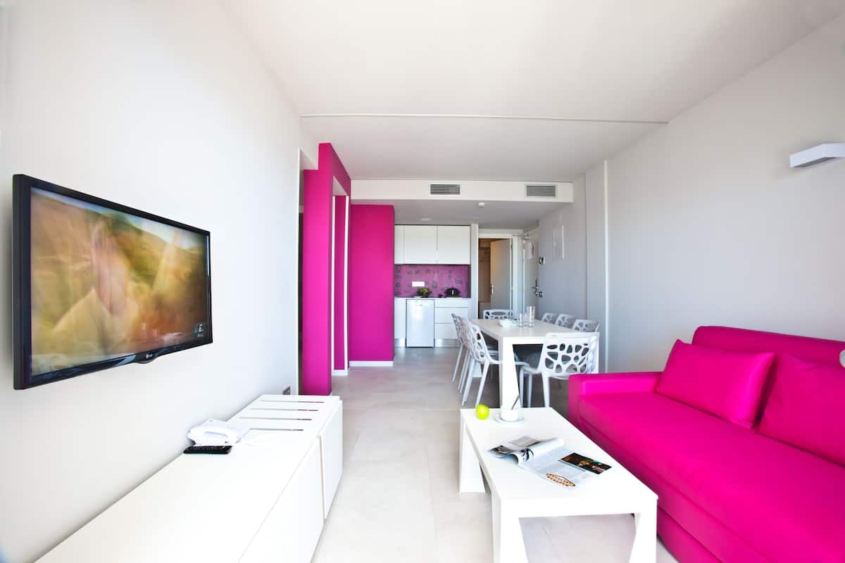 Una sala de estar moderna con un televisor de pantalla plana grande, un sofá rosa brillante, un comedor con muebles blancos y una cocina al fondo.