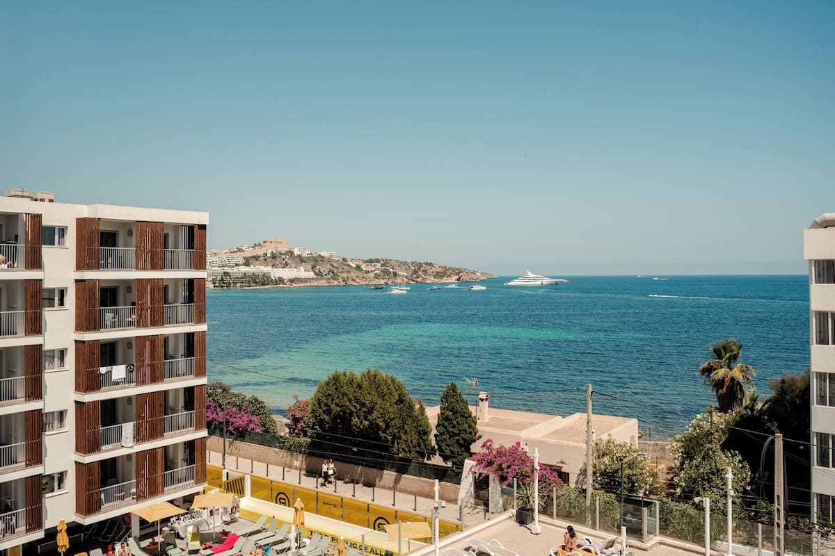 Un paisaje costero con edificios modernos, mar azul y una colina distante con construcciones.