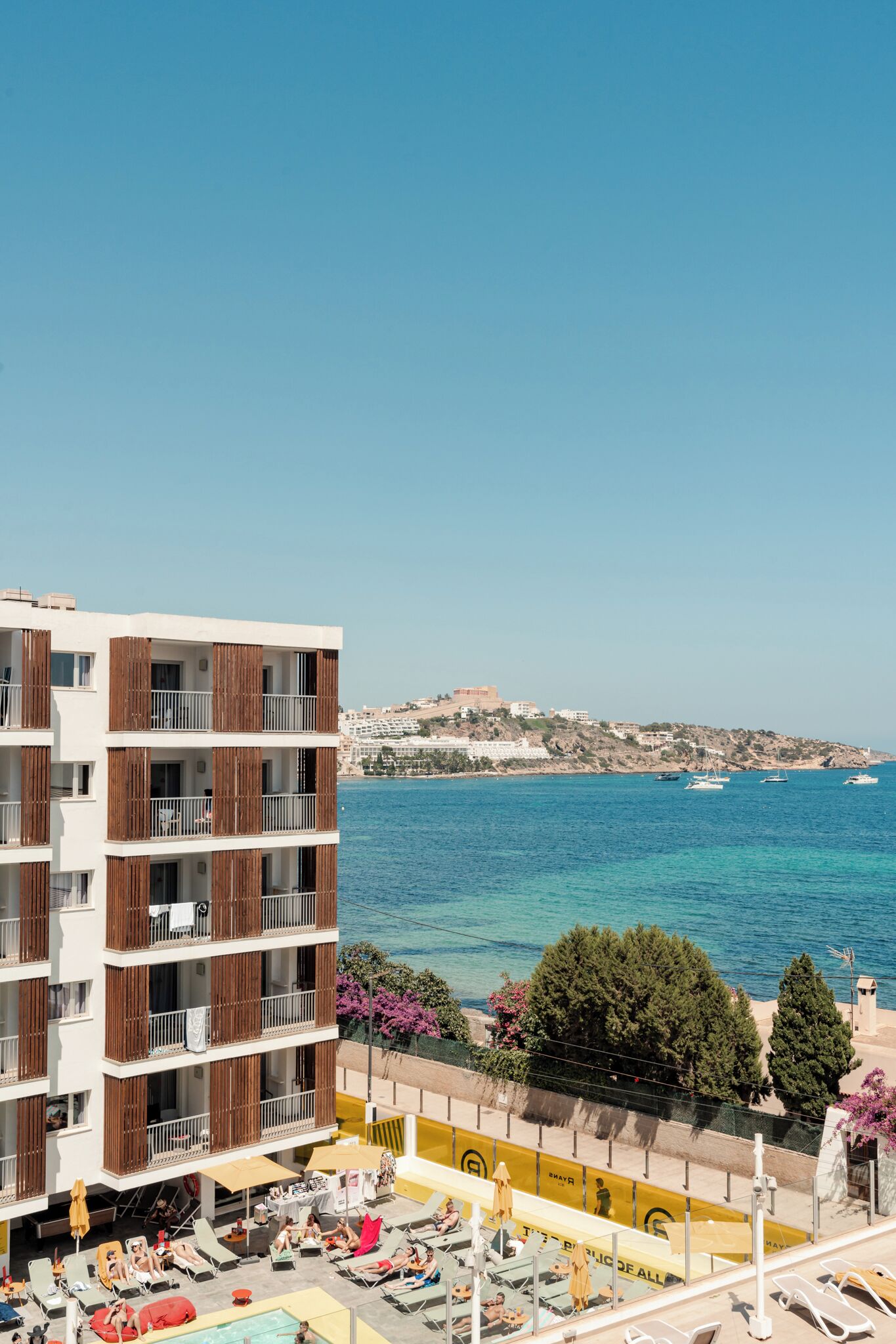 Ein mehrstöckiges Hotel mit Balkonen, einem Poolbereich und Blick auf das Meer und entfernte Gebäude.