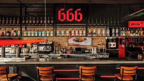Un bar con un llamativo letrero con el número '666', una variedad de botellas de licor, una máquina de café y tres taburetes de madera.