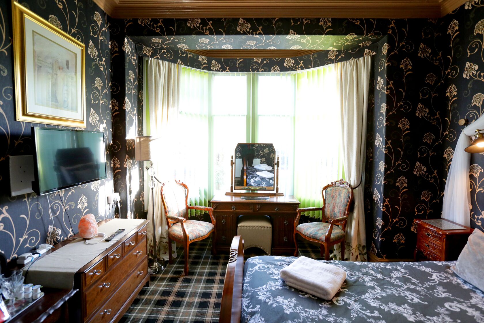 A room with a bed, two chairs, a desk, a television, and a painting.